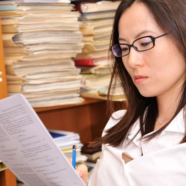 A woman with glasses reviews a document in a cluttered office.