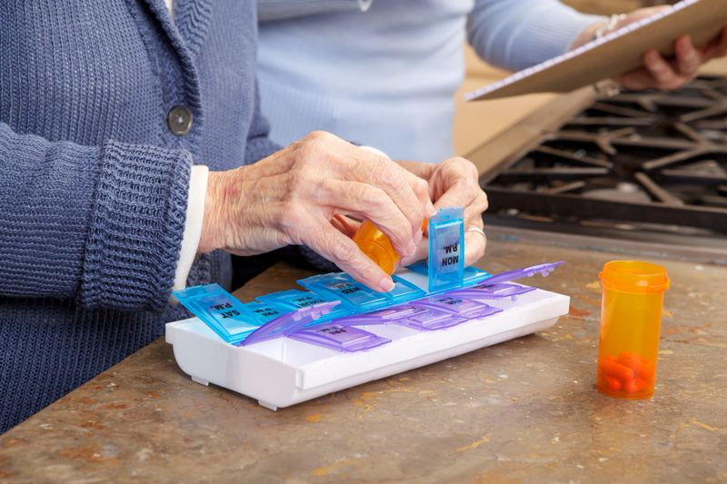 A close up view of an elderly woman adding pills to her organizer.  This organizer has compartments for both morning pills and evening pills each day.