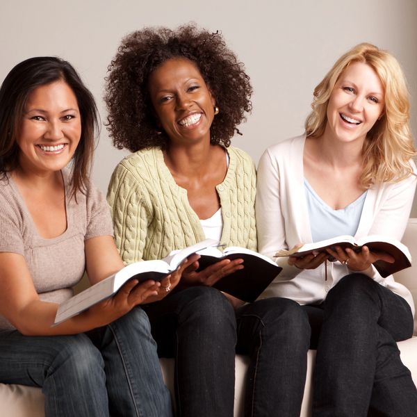 Three women smiling and reading books together indoors.