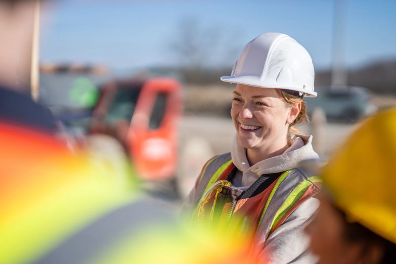 A small group of constructions workers meet with the forewoman to review the days work.  They are each wearing proper safety equipment and are listening attentively.