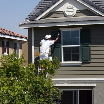 Painter on ladder painting house exterior near window and shutters.