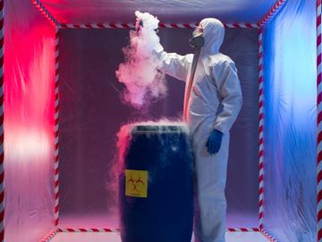 Person in hazmat suit handling a smoking biohazard barrel in a sealed room.