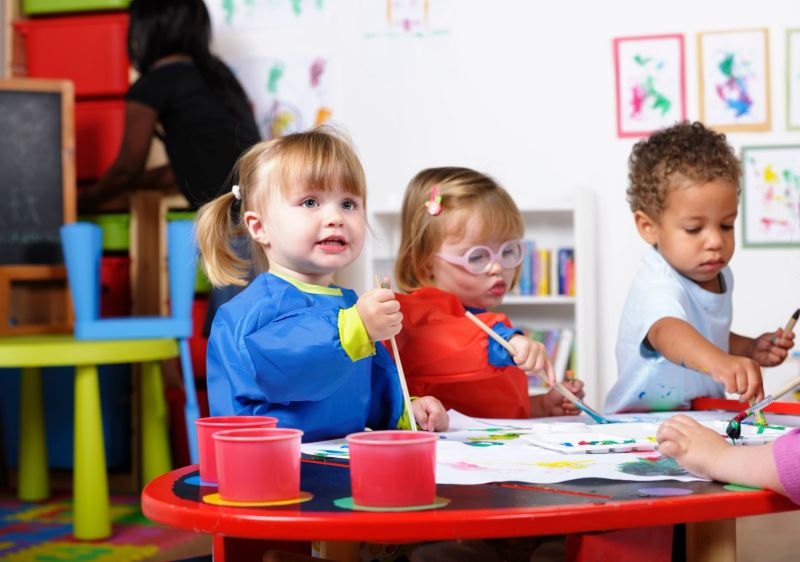 A portrait of a group of  toddlers painting whilt their carer tidies.