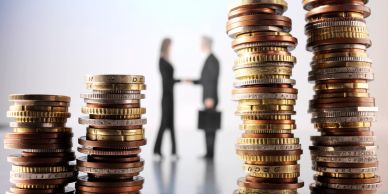 Stacks of coins with blurred business people shaking hands in background.