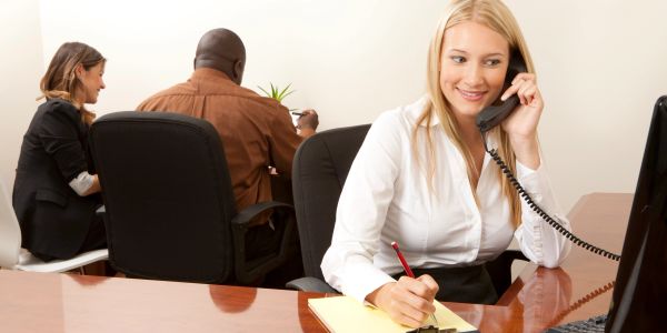 A smiling woman talks on the phone while taking notes at her desk in an office.
