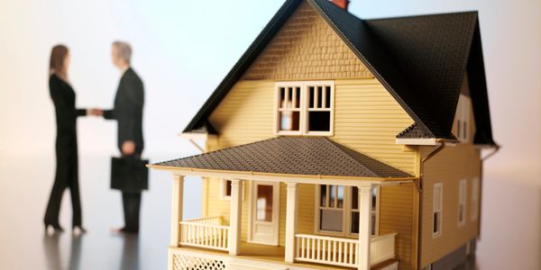Two professionals shaking hands behind a model house, symbolizing a real estate deal.