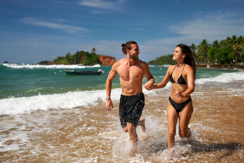 Joyful couple runs on tropical beach. Fit man and slender woman enjoy active vacation, sprinting by sea. Athletic duo practices beach running, promotes wellness. Excited pair in swimwear share fun.