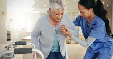 A nurse assists an elderly woman standing up with a walker nearby.