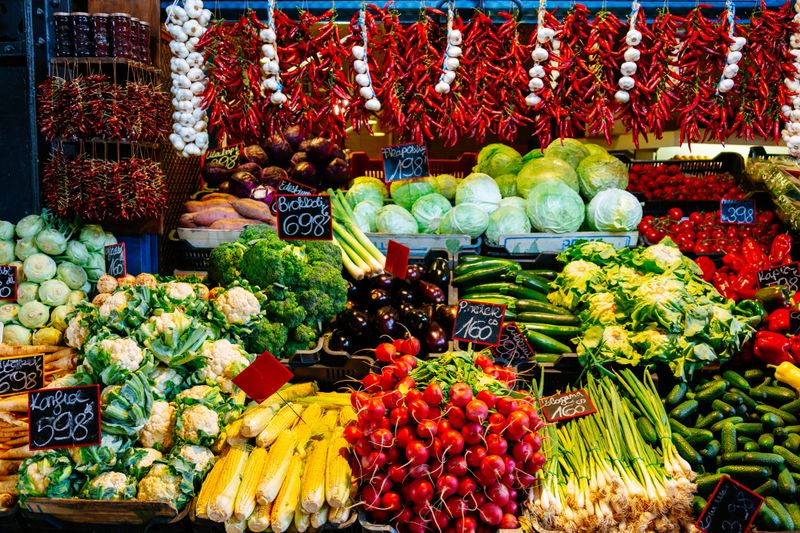 A market stall with an abundance of fresh vegetables and fruits. The stall is filled with a variety of colorful produce such as red peppers, green cabbage, yellow corn, and purple eggplants. The produce is arranged in neat piles and rows, with small price tags scattered throughout.