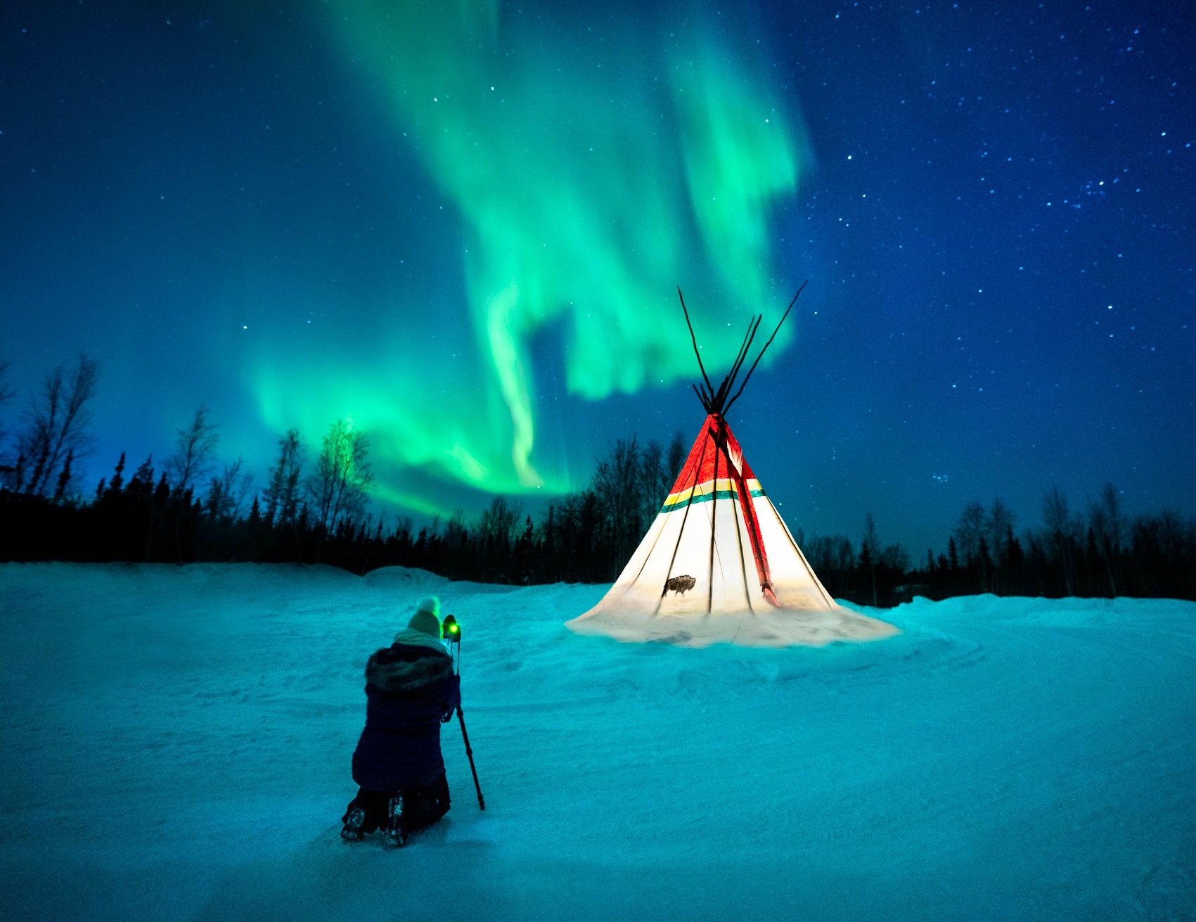 A person photographing a tipi under the Northern Lights at night.