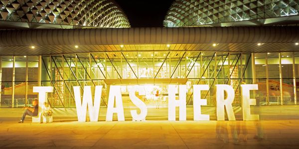 Large illuminated letters spell "I WAS HERE" at night in front of a modern building.