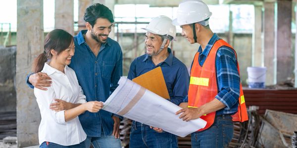 Four construction professionals discussing blueprints on site, two wearing helmets and one in a safety vest.