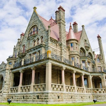 Large historic stone mansion with towers and wrap-around porch under a partly cloudy sky.