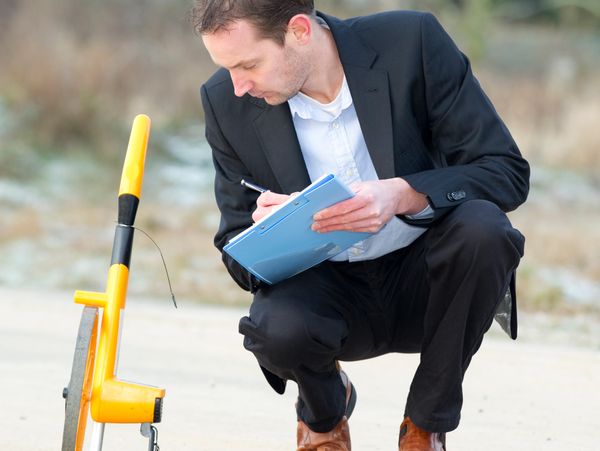 Man in a suit measuring and recording data on a clipboard outdoors.