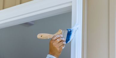 A hand using a paintbrush to apply white paint to a window frame, captured during a painting project.