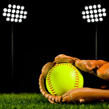 A softball nestled in a glove on grass under stadium lights.
