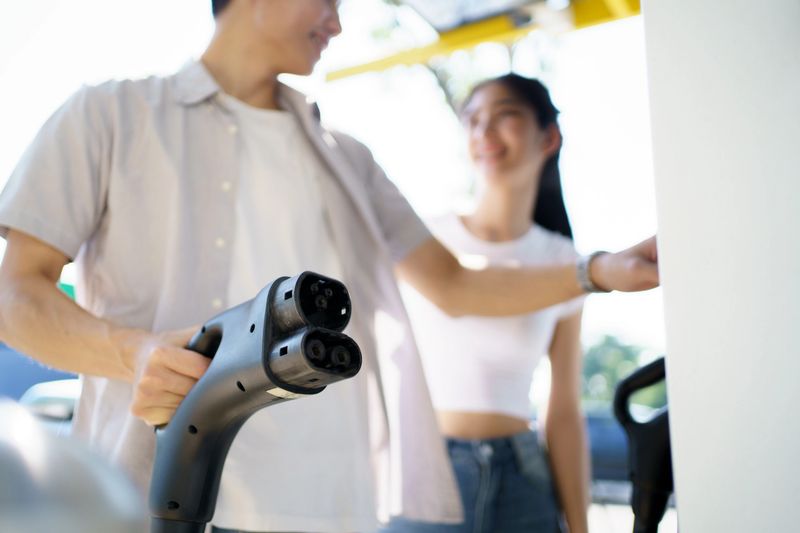 Happy Asian lovely couple using EV charging application on smartphone or mobile phone to access to the charging station. Man plug-in the DC CCS type-2 charging connector to the electric vehicle, meanwhile his girlfriend standing beside him at public charging station.