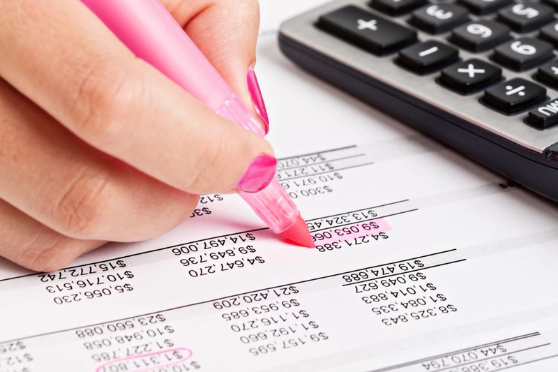 "A woman's hand checks a financial document, highlighting a figure in pink, as a calculator stands by. Could be an audit or home or office finances being checked so carefully."