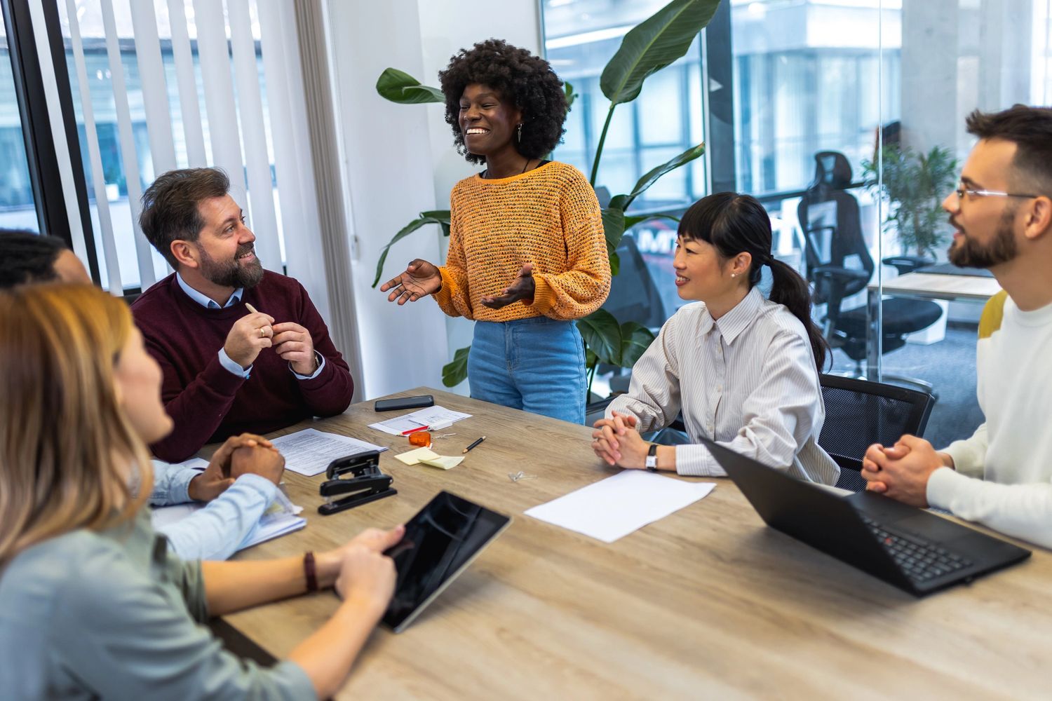 A woman in an orange sweater leads a lively meeting with engaged colleagues around a table.