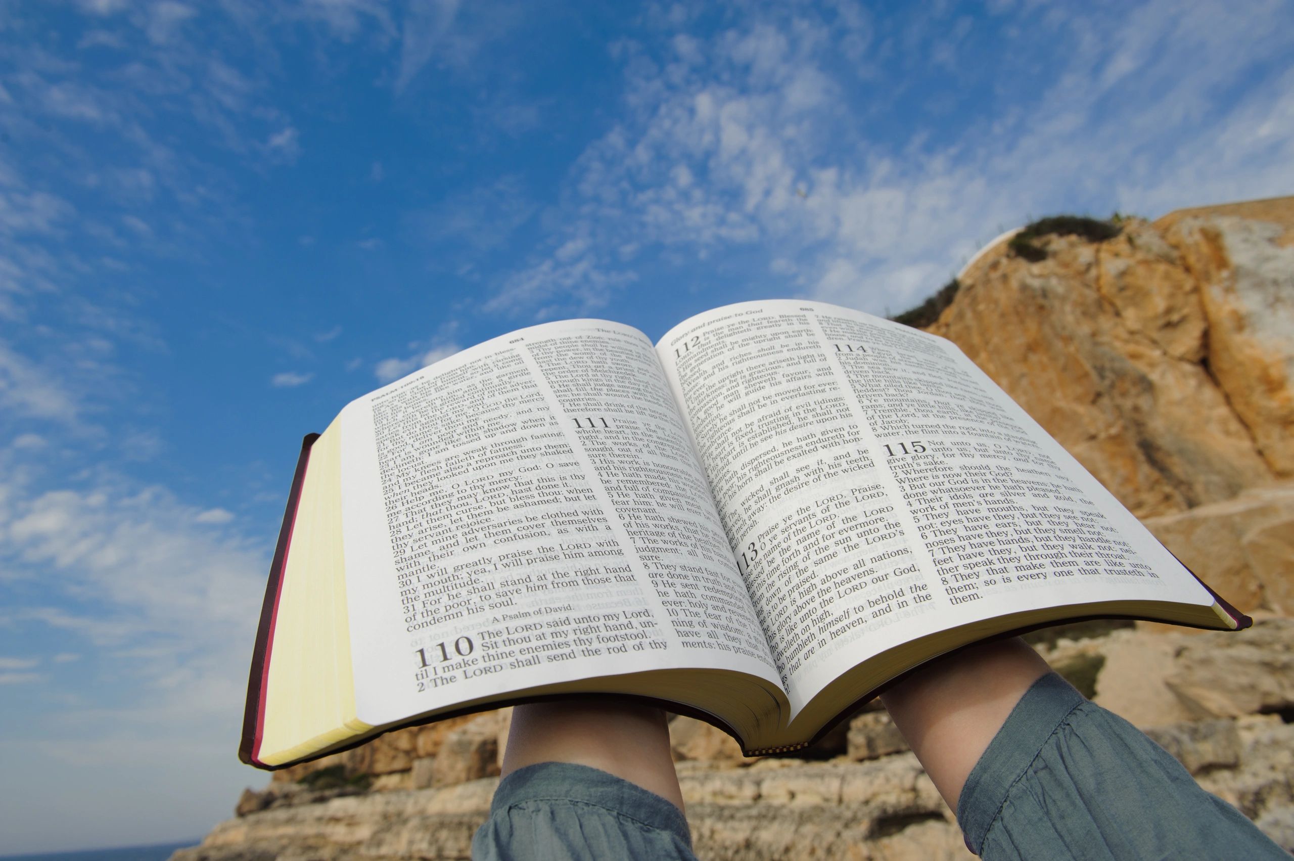 Person holding an open Bible against a blue sky and rocky background.