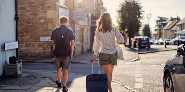 Two people walking down a sunny street, one pulling a suitcase.