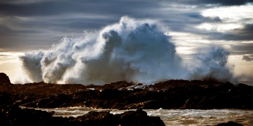 Powerful ocean waves crashing against dark rocky shore under a moody sky.