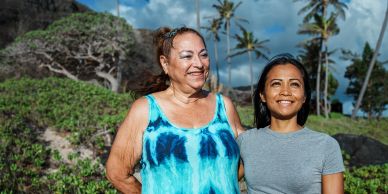 Two women smiling outdoors with trees and blue sky.