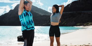 Two women stretching on a sunny beach with mountains in the background.