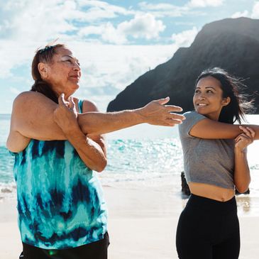 Two women stretching arms on a sunny beach with mountains and ocean in the background.