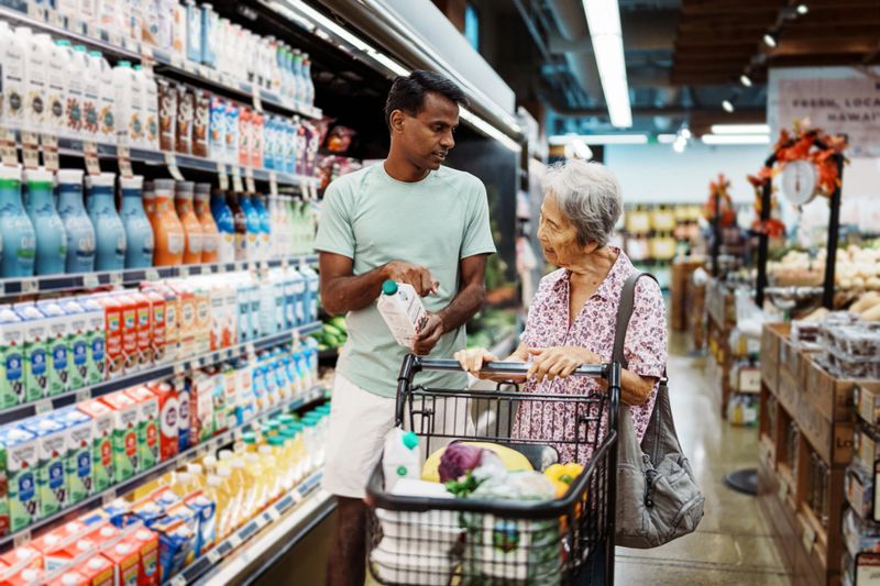 An elderly Korean woman and her caregiver review the nutrition information on a product while shopping in the refrigerated dairy section of an organic grocery store. The home health care provider is working for a community outreach program aimed to provide support and nutrition guidance to senior adults.