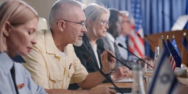 Diverse officials speak at a conference table with microphones and flags.