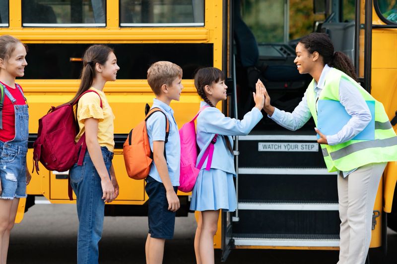 Kids boarding school bus receiving joyful high five from their smiling black teacher, happy children greeting attendant driver woman in uniform, ready for journey together, side view