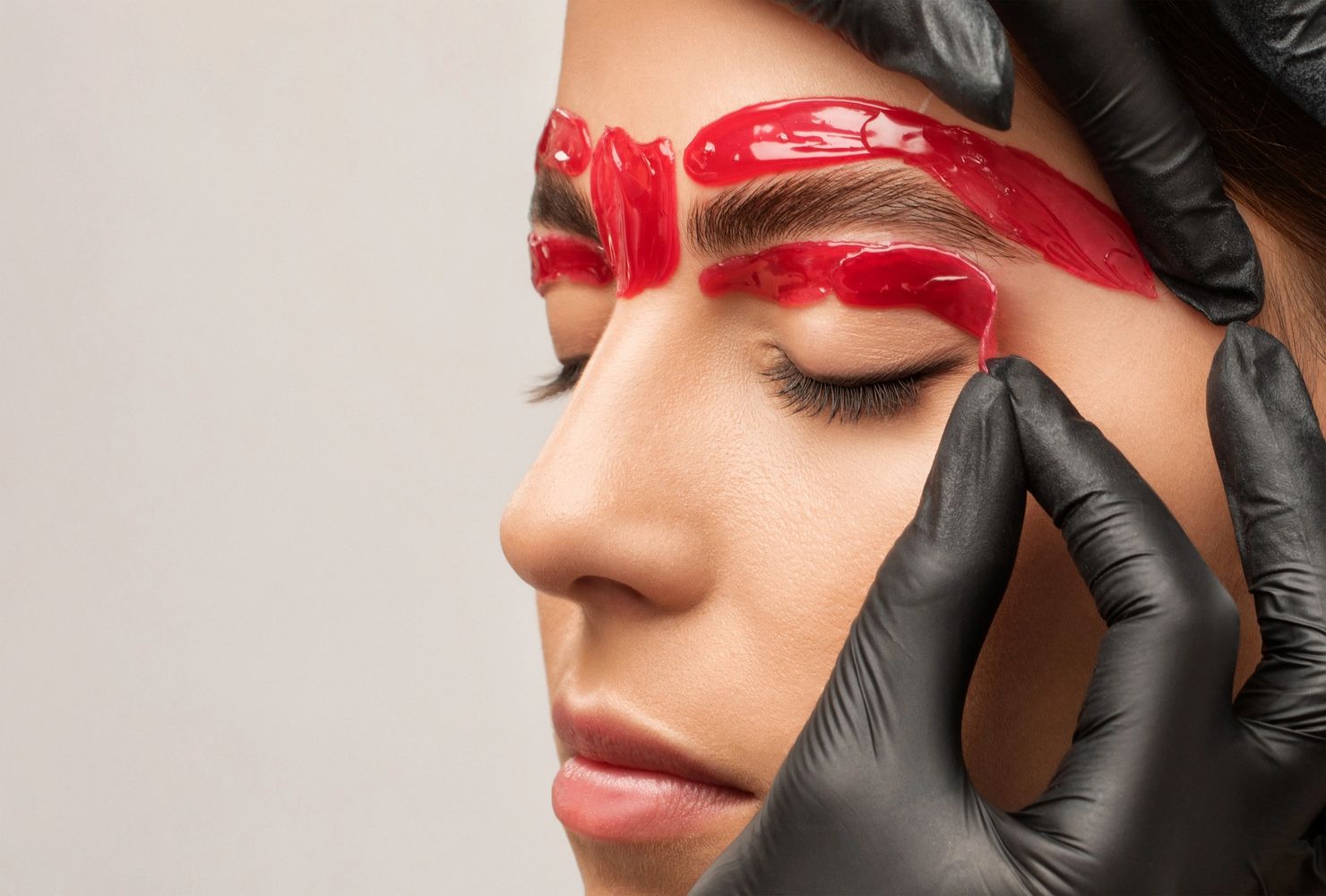 Close-up of eyebrow waxing with red wax being peeled off by gloved hands.