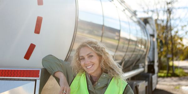 Smiling woman in safety vest leaning on a tanker truck outdoors.