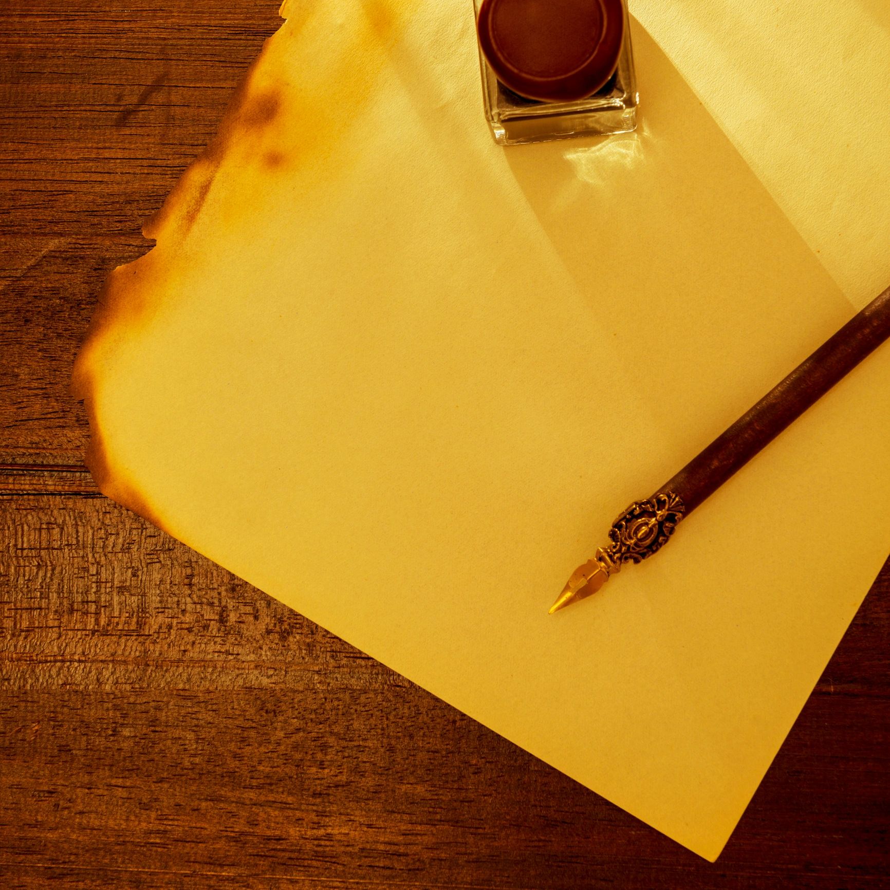 Burnt-edged parchment with an ink bottle and ornate dip pen on wood.