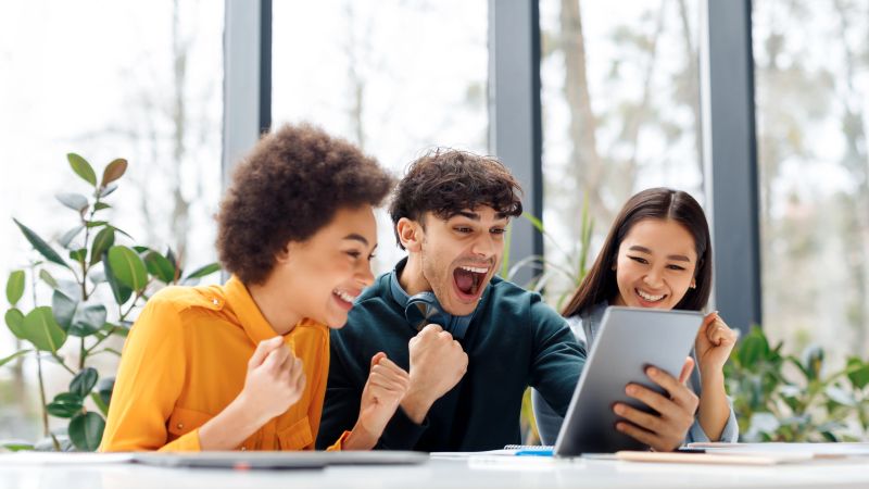 Overjoyed multiracial classmates looking at digital tablet screen, shouting yes and clenching fists in excitement, sitting in classroom, panorama with free space