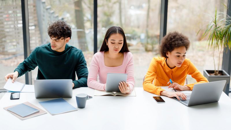Multiracial students sitting in university audience, using various gadgets, learning for exams, multiracial guy and ladies making online research indoors