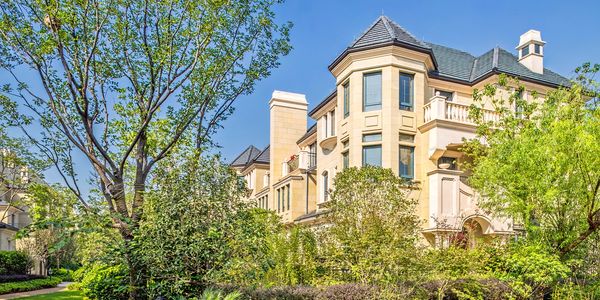 Elegant beige mansion surrounded by lush greenery under a clear blue sky.