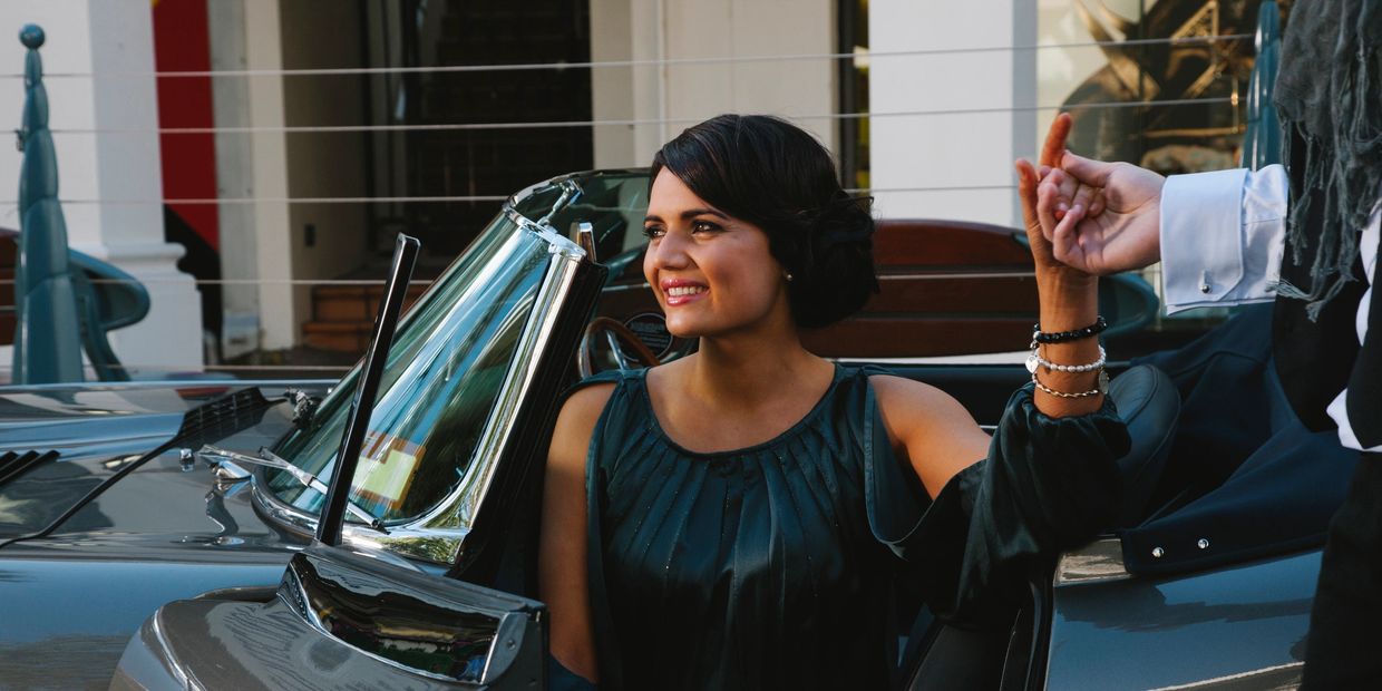 A smiling woman in a vintage car holding hands with someone outside the frame.