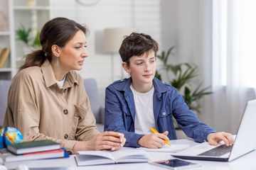 A woman helps a boy with homework using a laptop.
