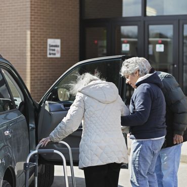 An elderly woman with a walker is helped into a car by two people.