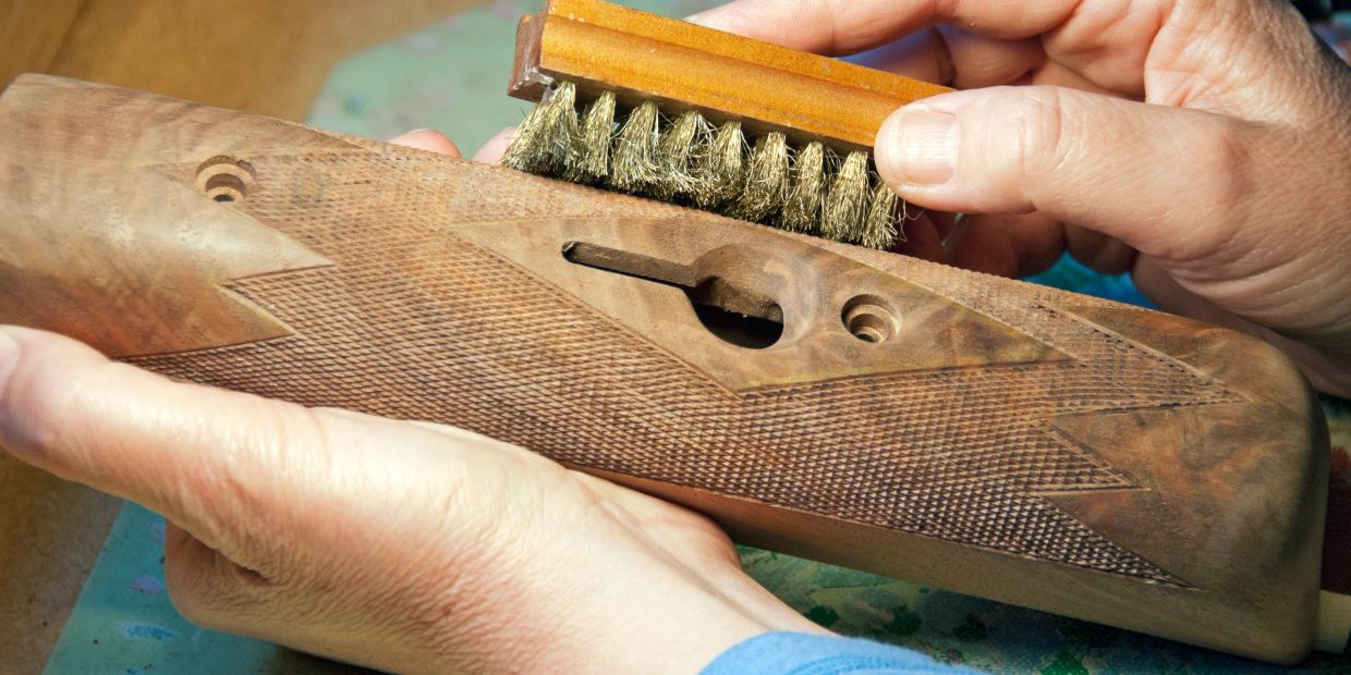 Hands cleaning a wooden gun stock with a brush.