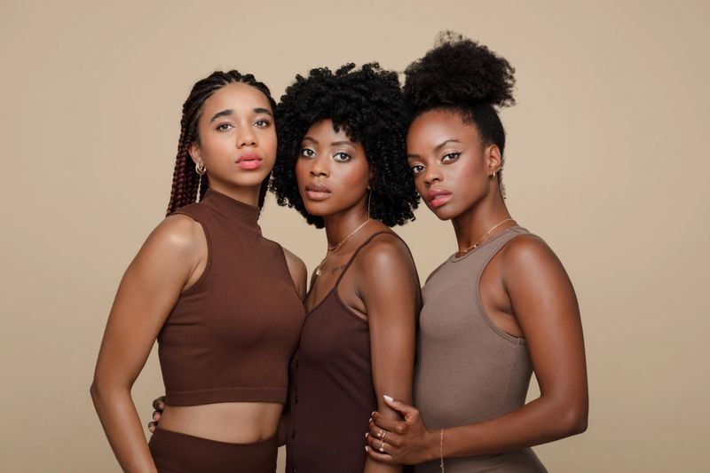 Three beautiful young women wearing brown clothes standing against beige background, embracing and looking at camera.