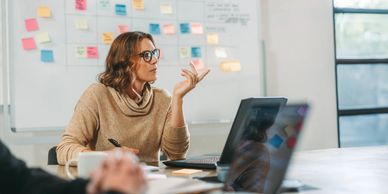Woman in glasses attentively listens during a meeting with a laptop and sticky notes board.