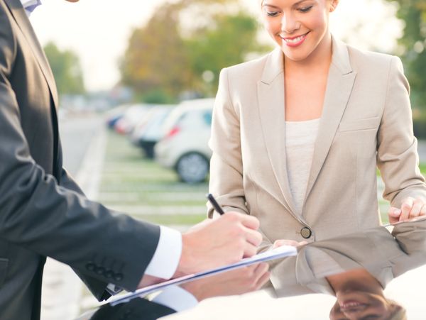 A woman in a beige suit smiles while a man signs a document outdoors.