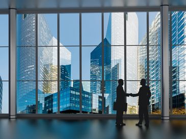 Two businesspeople shake hands in a modern office with city skyscrapers outside.