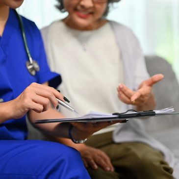 A healthcare professional in blue scrubs reviews documents with a patient on a couch.