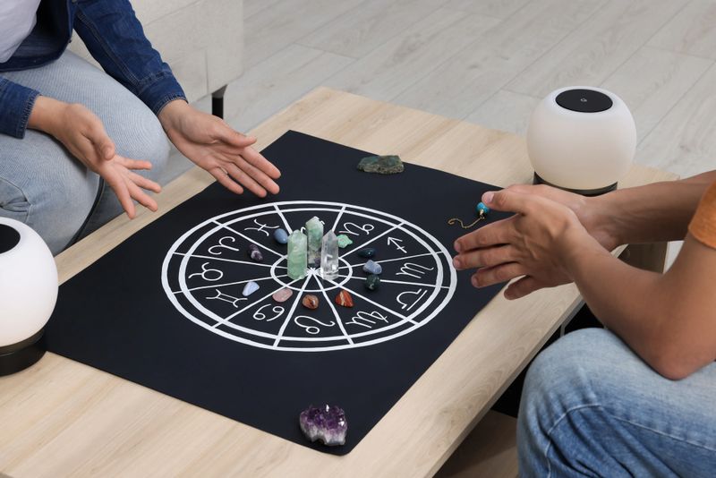 Astrologer predicting client's future with zodiac wheel, stones and crystals at wooden table indoors, closeup