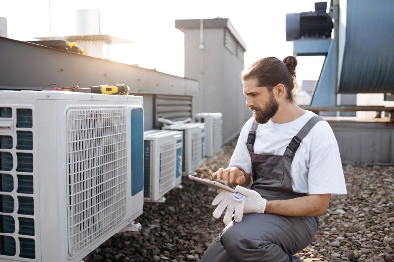 Focused master wearing gray overalls holding wireless tablet while inspecting air conditioner on plant. Caucasian qualified craftsman crouching and comparing information with device outdoors.