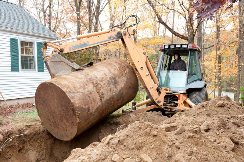 A man sitting in an excavator removes an old, rusty underground oil tank from a residential yard.